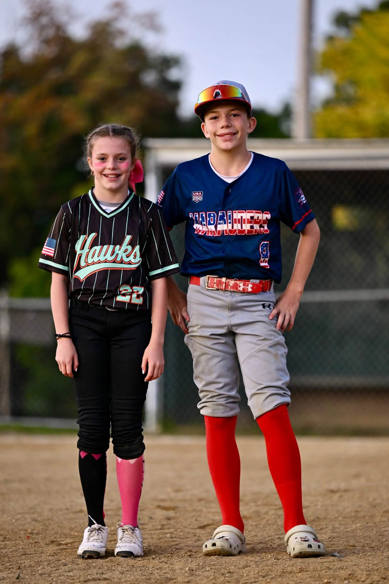 Reese in her softball uniform and Cal in his baseball uniform standing side by side on an infield.